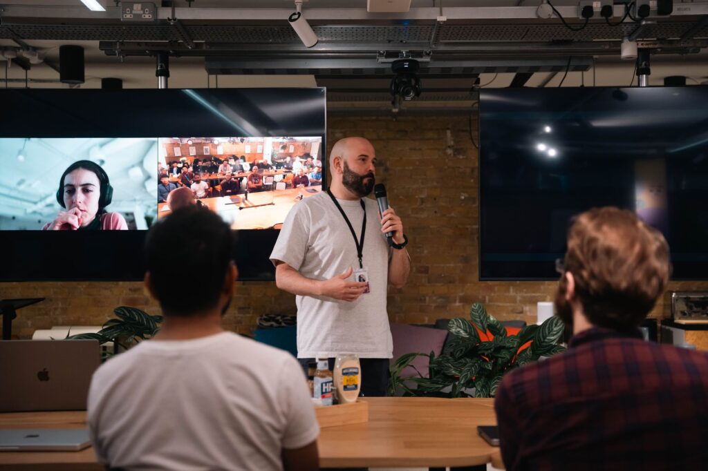 Speaker presenting in a modern office with audience and virtual call on screens.