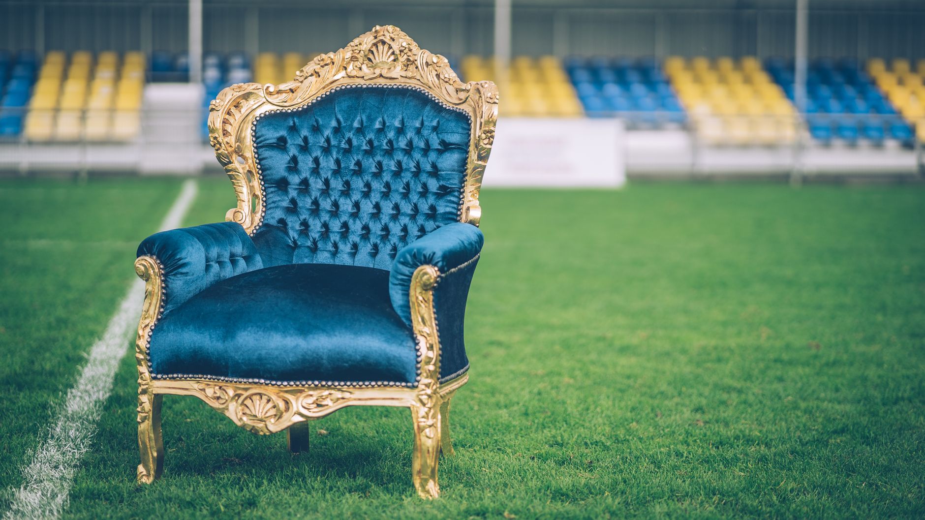 A luxurious blue velvet and gold armchair sits prominently on a sports field.