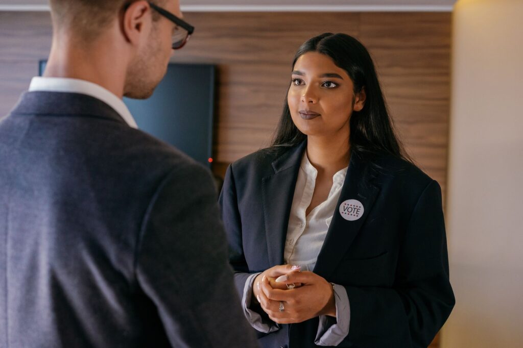 A female candidate in a political discussion, wearing a vote badge during an election campaign indoors.