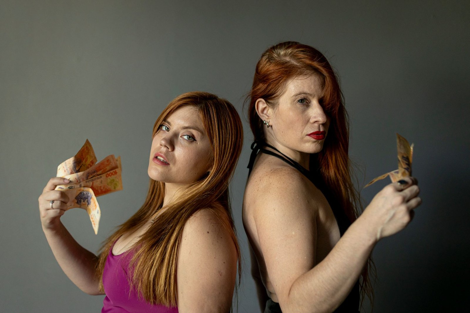Portrait of two women displaying Argentine pesos. Shot indoors with a confident look.
