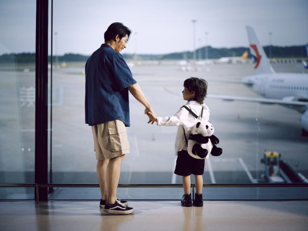 A father and child holding hands, gazing at planes through an airport window.