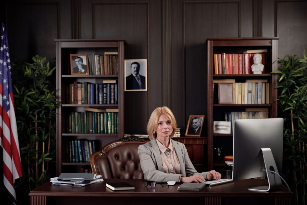 A businesswoman sits confidently at her desk in an office setting with a bookshelf and American flag.