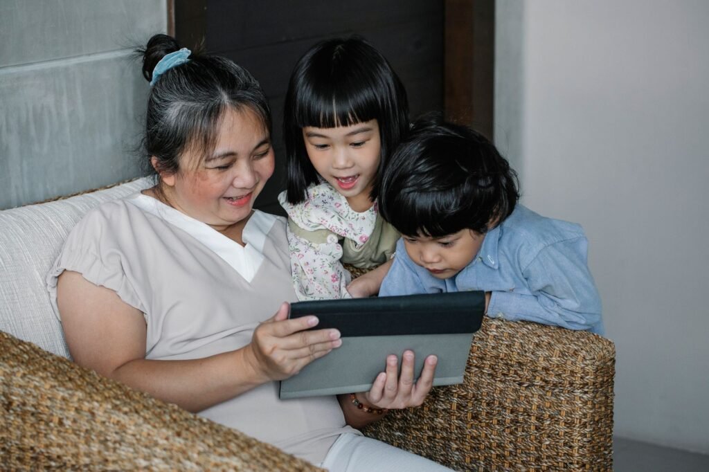 Happy Asian babysitter sitting on armchair with kids and browsing tablet in cozy apartment