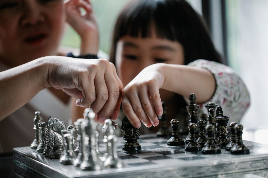 Thoughtful Asian girl playing stylish chess with babysitter against window in light room in daytime