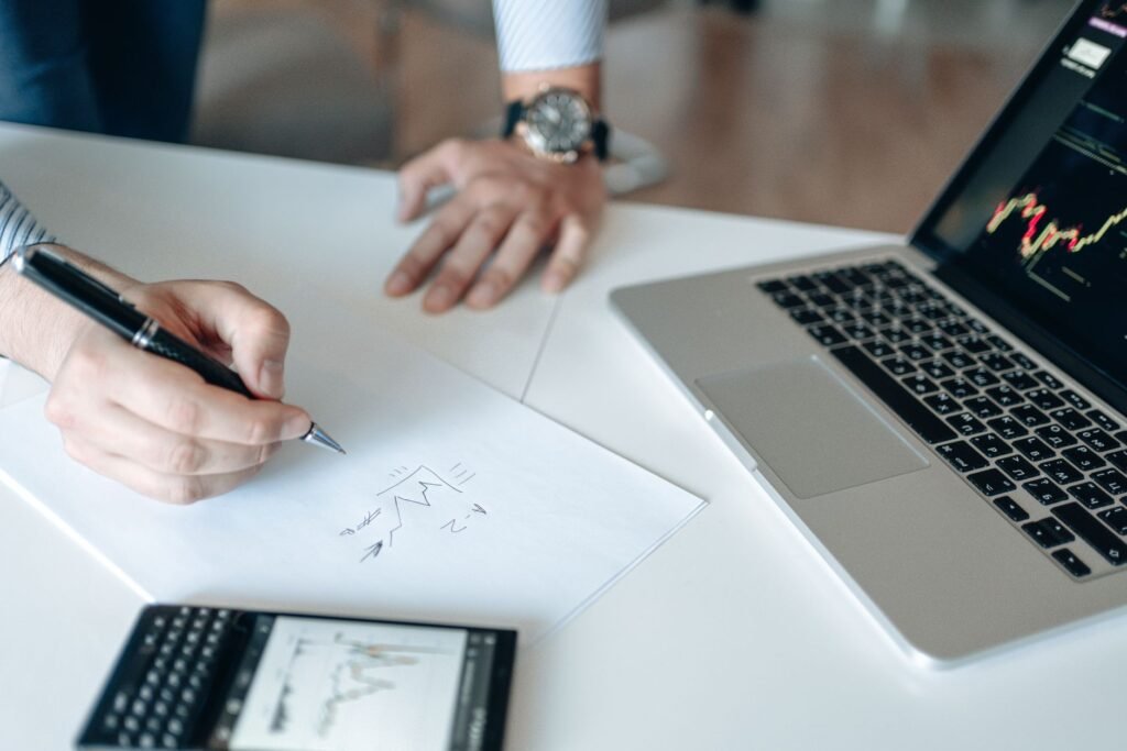 Close-up of hands drawing stock trends on paper with laptop and smartphone on desk.