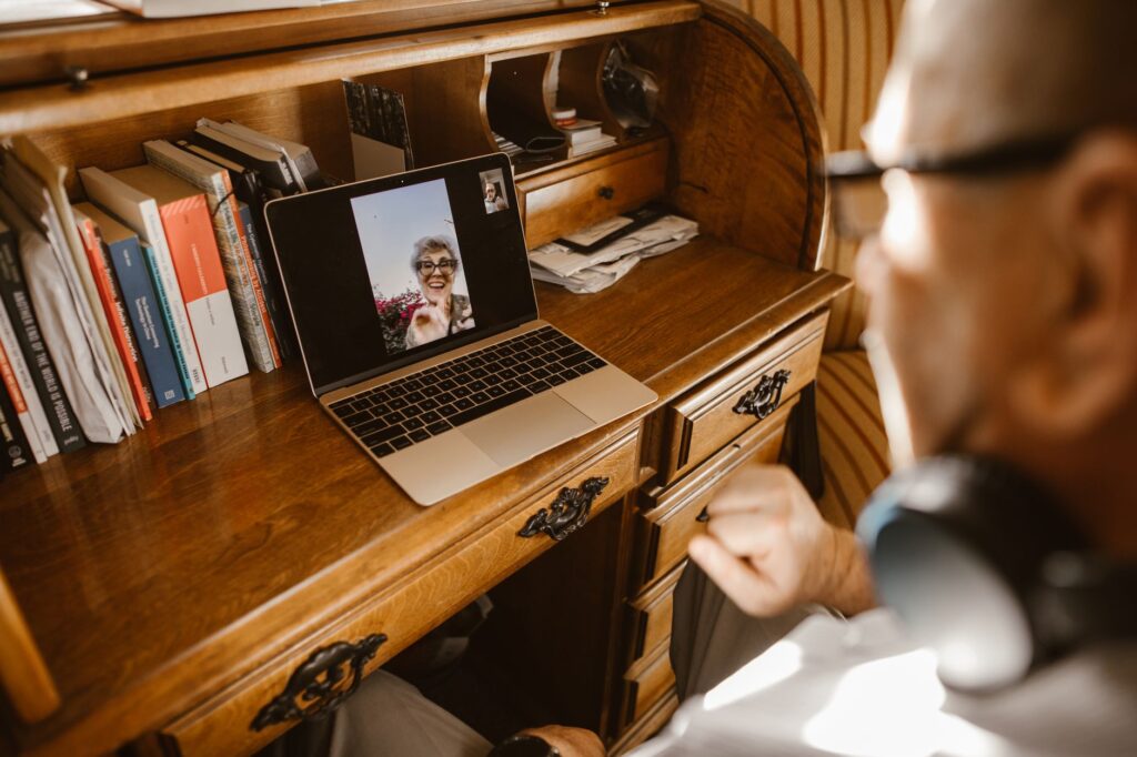 An elderly man video calling a woman using a laptop at a wooden desk.