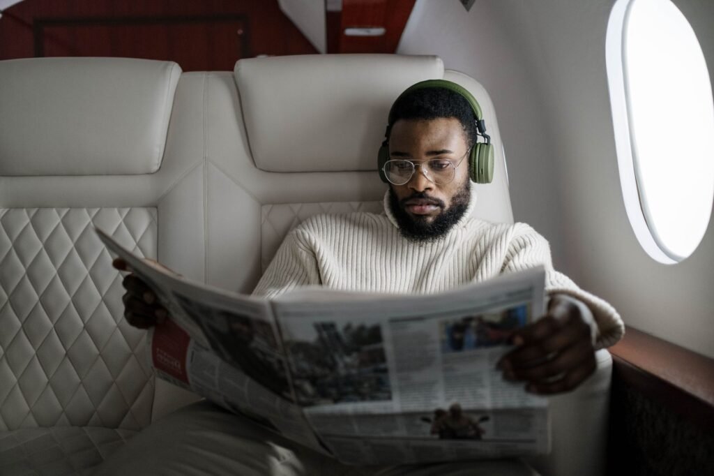 Man wearing headphones reads a newspaper while seated on an airplane during the daytime.