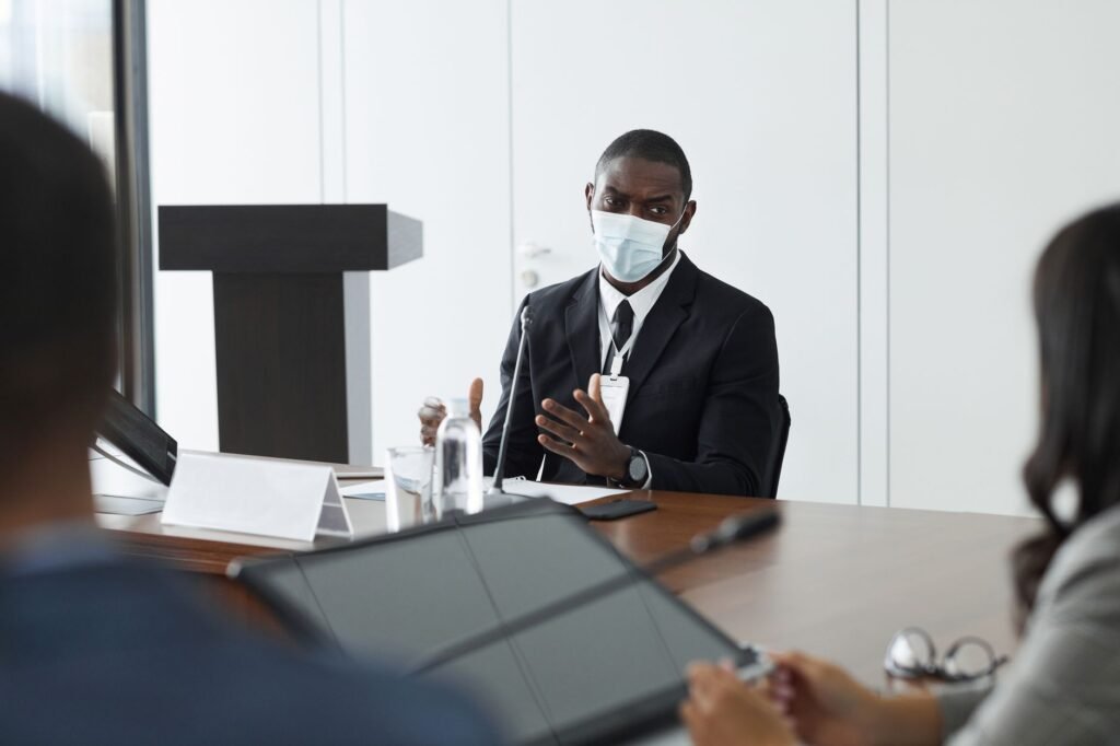 A masked professional speaks at a formal meeting, emphasizing safety and communication.
