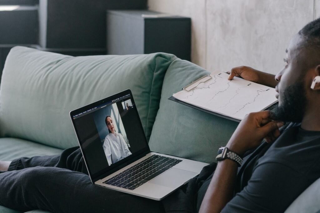 A man holding a clipboard while on a video call from a laptop, seated on a sofa.