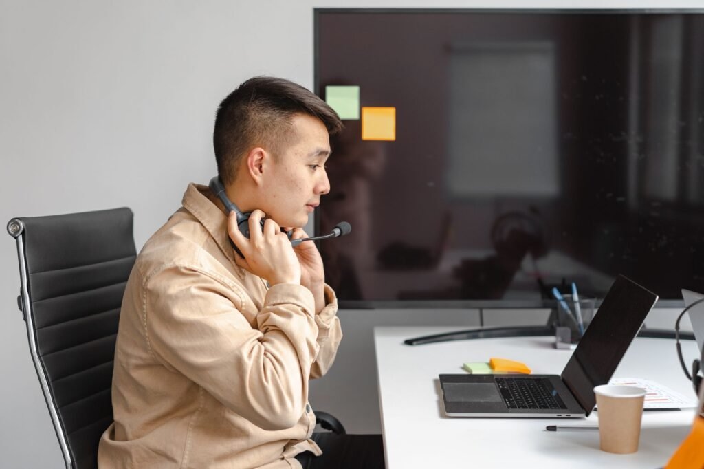 Man working with laptop in office setting, wearing a headset for an online meeting.