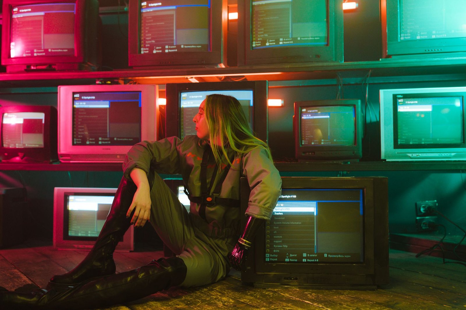 A woman with a prosthetic hand sits in a neon-lit room surrounded by retro TVs.
