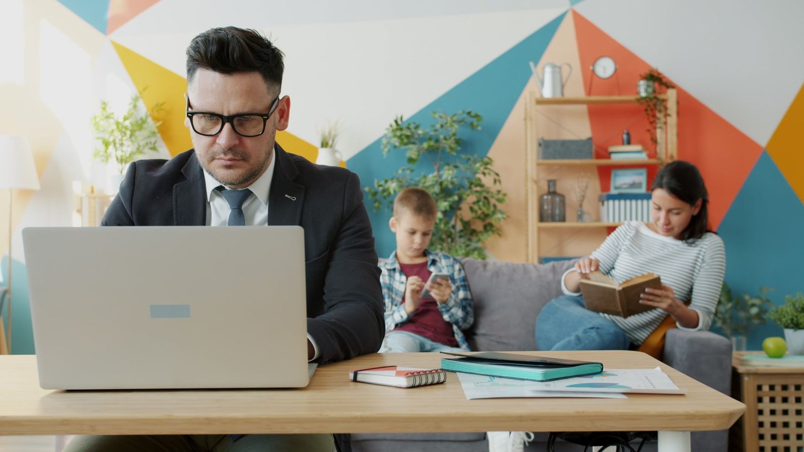 A man working on a laptop at home with family relaxing in the background.