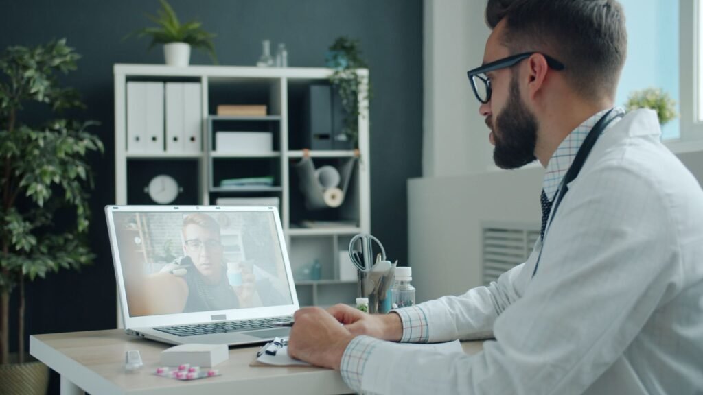 Doctor in white coat video calling a patient on a laptop in a modern office setting.
