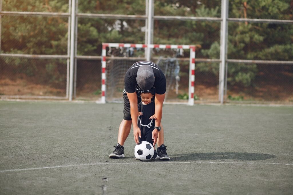 Full body anonymous father in black casual wear and toddler son walking with football ball on green sports ground on sunny day