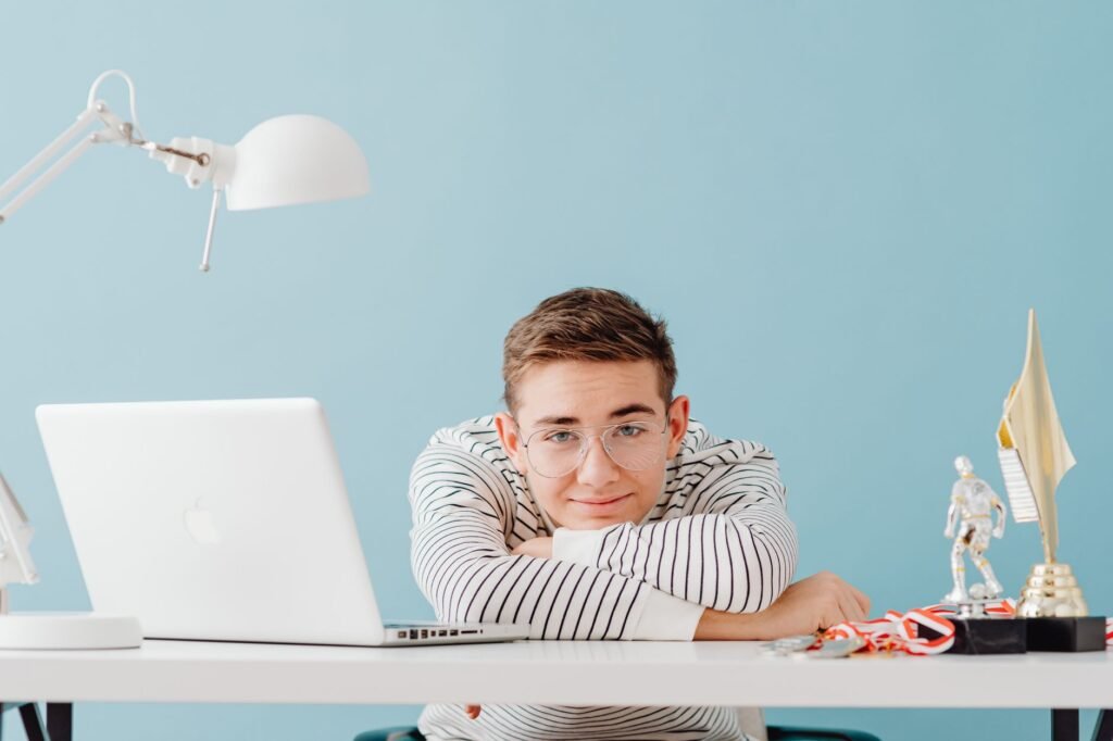 Young man with glasses leaning on a desk with a laptop and trophies, against a teal background.