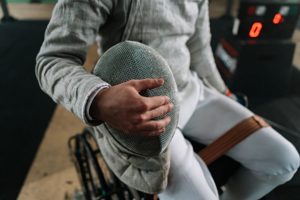 Fencer in wheelchair holding mask, ready for competition in indoor gym setting.